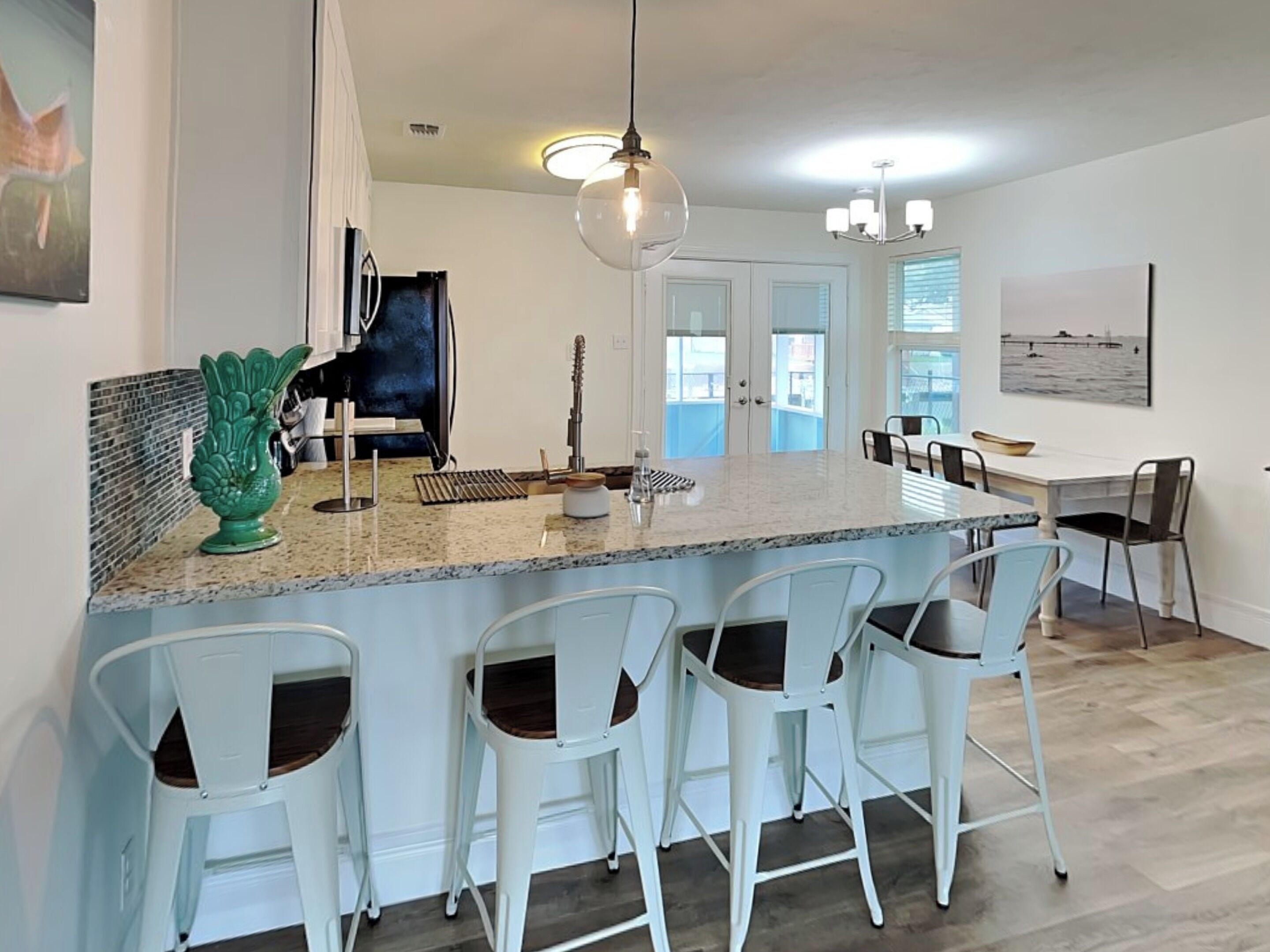 A modern kitchen with a granite countertop, white bar stools, and a dining area with a view of the ocean.