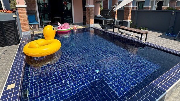 Una piscina con mosaicos azules, rodeada por una casa de fachada rosa y tejado de baldosas.
