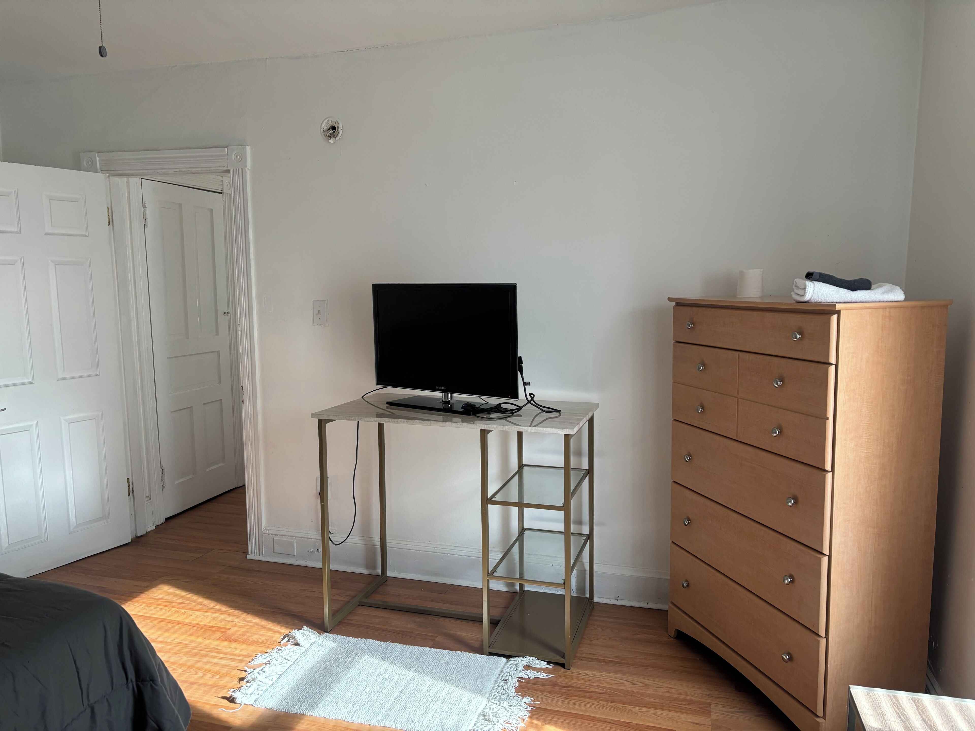 A bedroom with a bed, a desk with a TV, a dresser, and a door.
