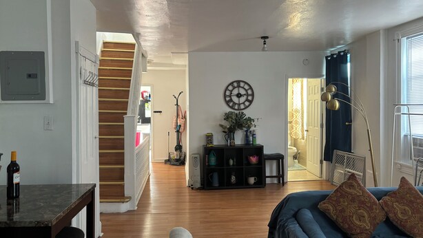 A spacious living room with a staircase, a coffee table, a sofa with patterned cushions, and a wall clock.
