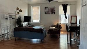 A living room with a blue sofa, a small table, a TV, and a ceiling fan. There are two windows with blinds, a ladder, and a plant.