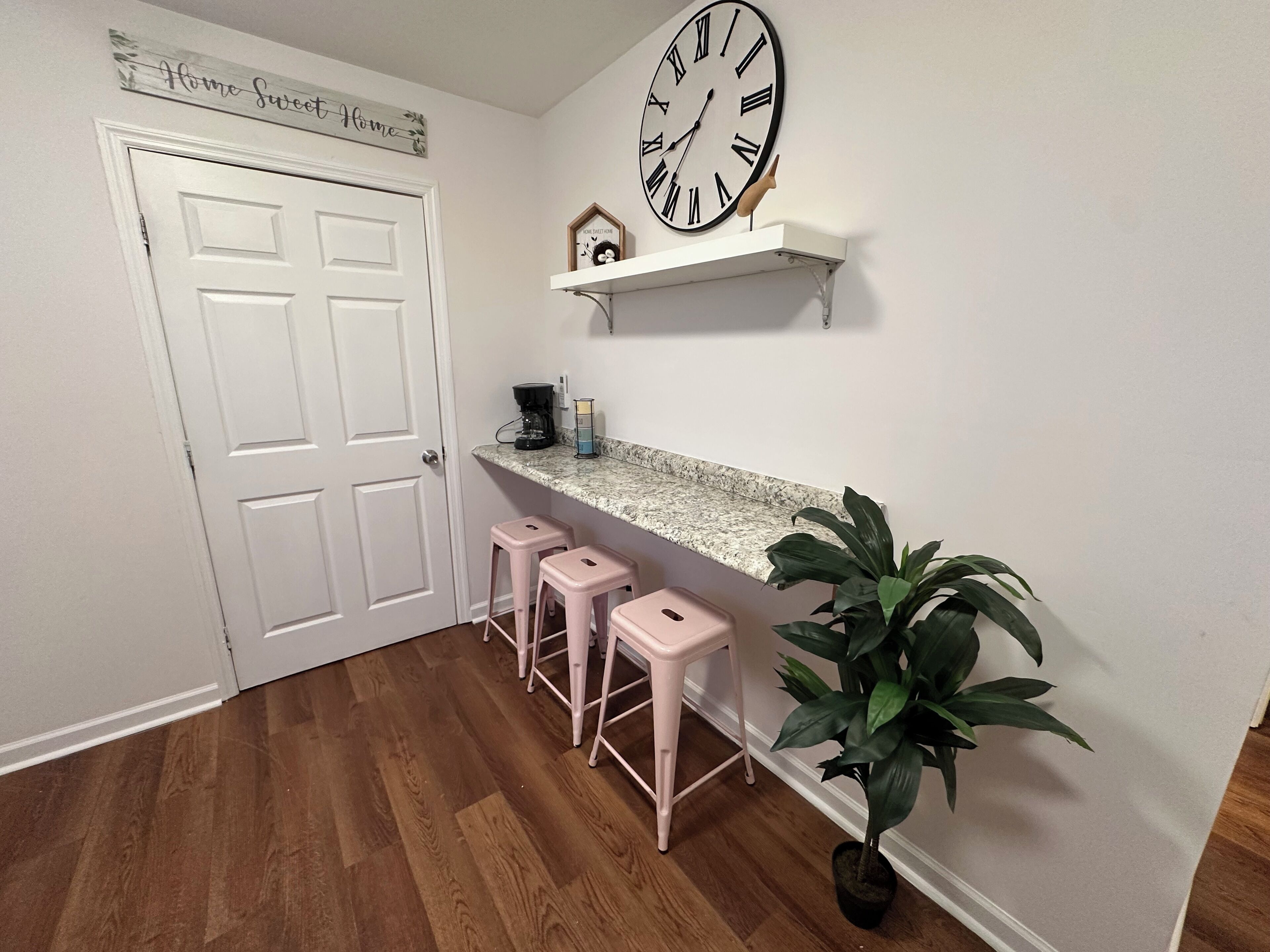 A kitchen with a white door, a large wall clock, a coffee maker, a shelf with decorative items, and three pink barstools.