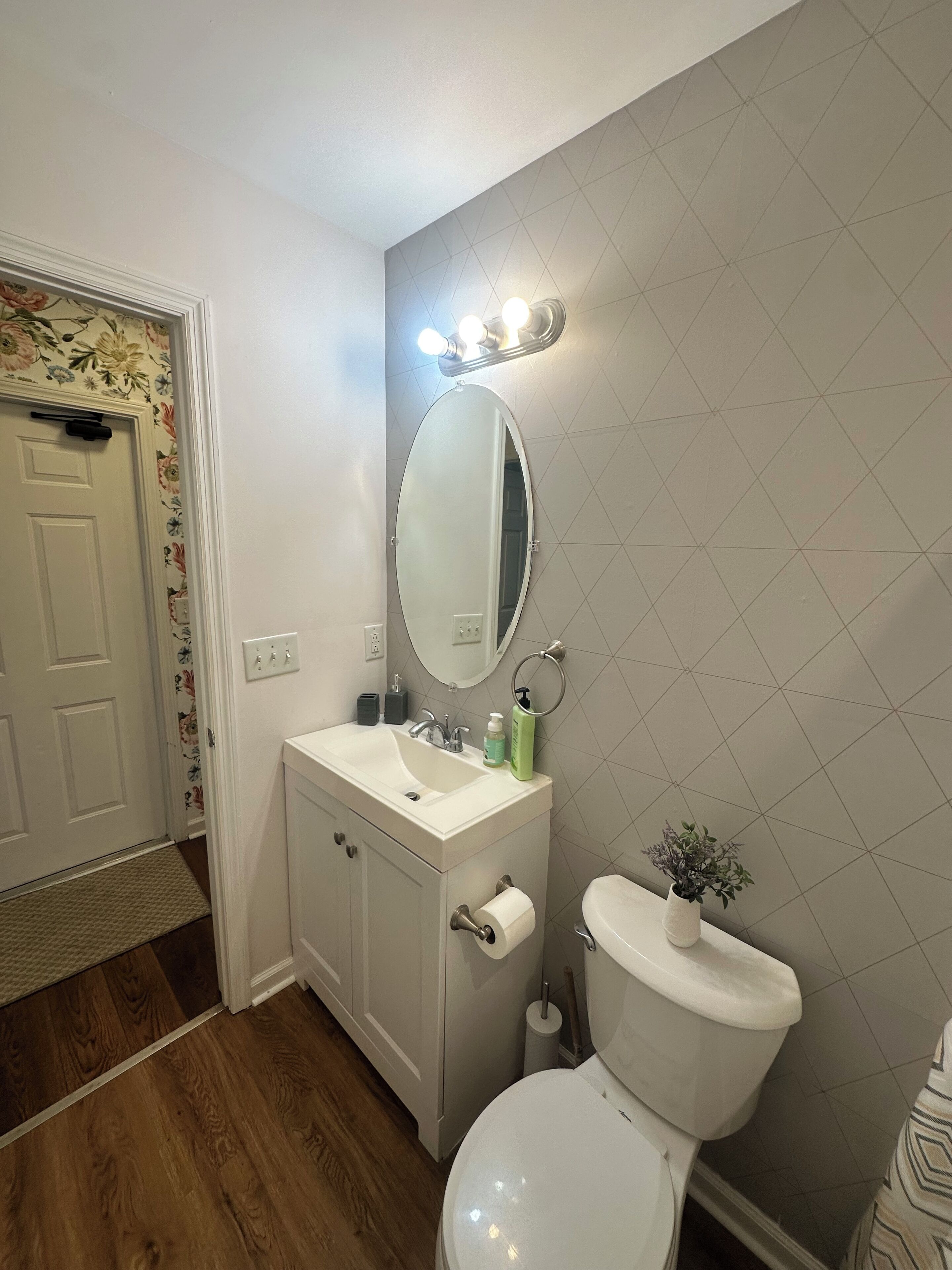 Ein Badezimmer mit Waschbecken, Spiegel und Toilette. An der linken Wand ist ein Blumenmuster, an der rechten eine geometrische Wandgestaltung.