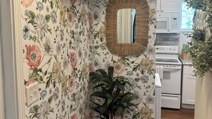 A hallway with floral wallpaper, a potted plant, an air conditioning unit, and a kitchen area with white appliances.