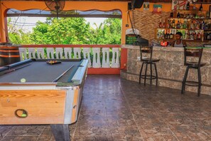 Un tavolo da biliardo con una palla verde, un'area bar con sgabelli e vista sul verde circostante.