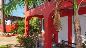 Un bâtiment rouge avec une porte blanche, un escalier et un petit jardin agrémenté de plantes et d’un banc.