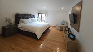 A bedroom with a large bed, a bedside table, a nightstand, a TV mounted on the wall, and a window with a view.