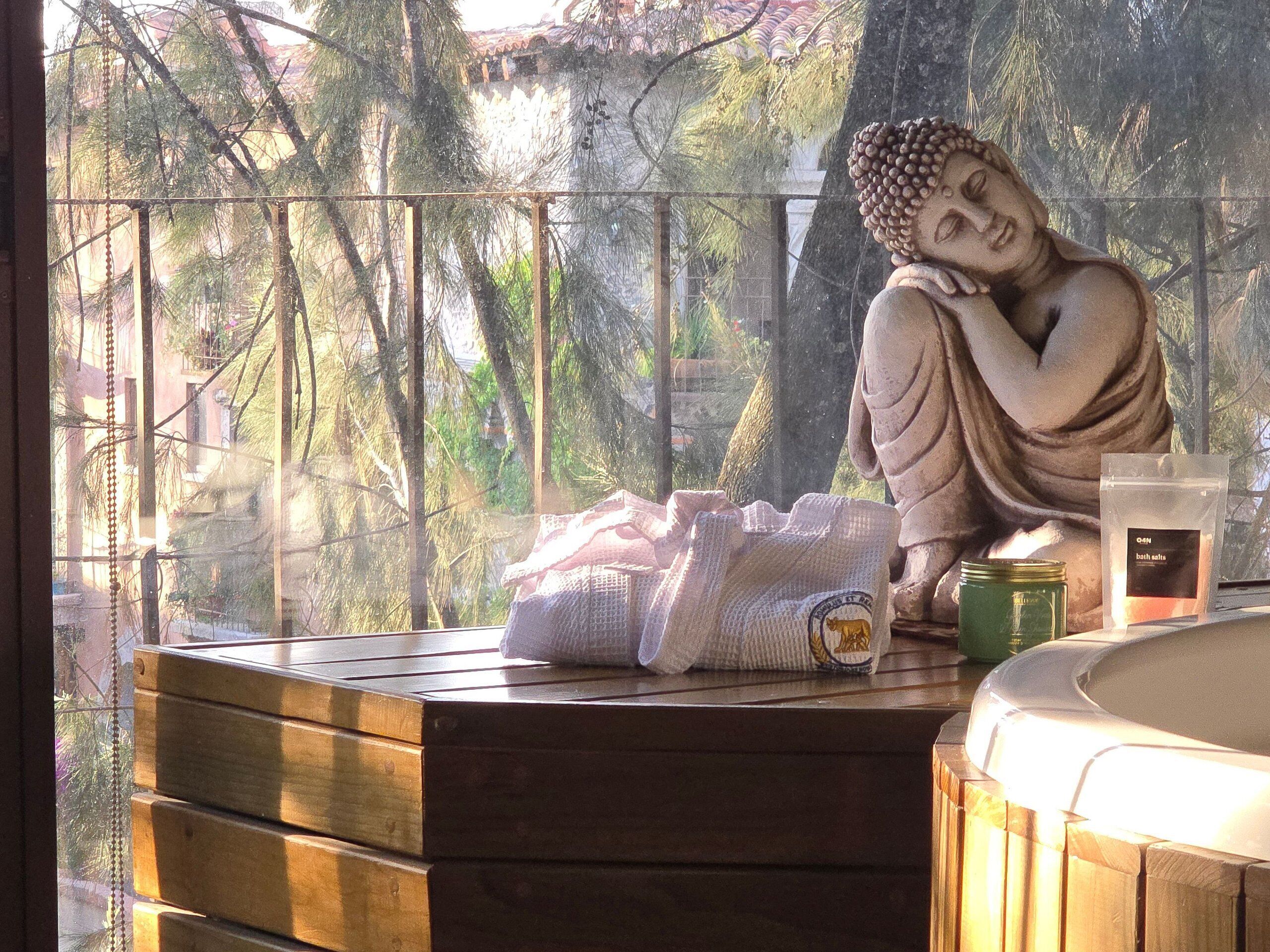 A statue of Buddha on a wooden bench with a towel and a candle, overlooking a balcony with trees.