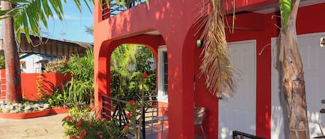 A red building with a white door, a staircase, and a small garden area with plants and a bench.