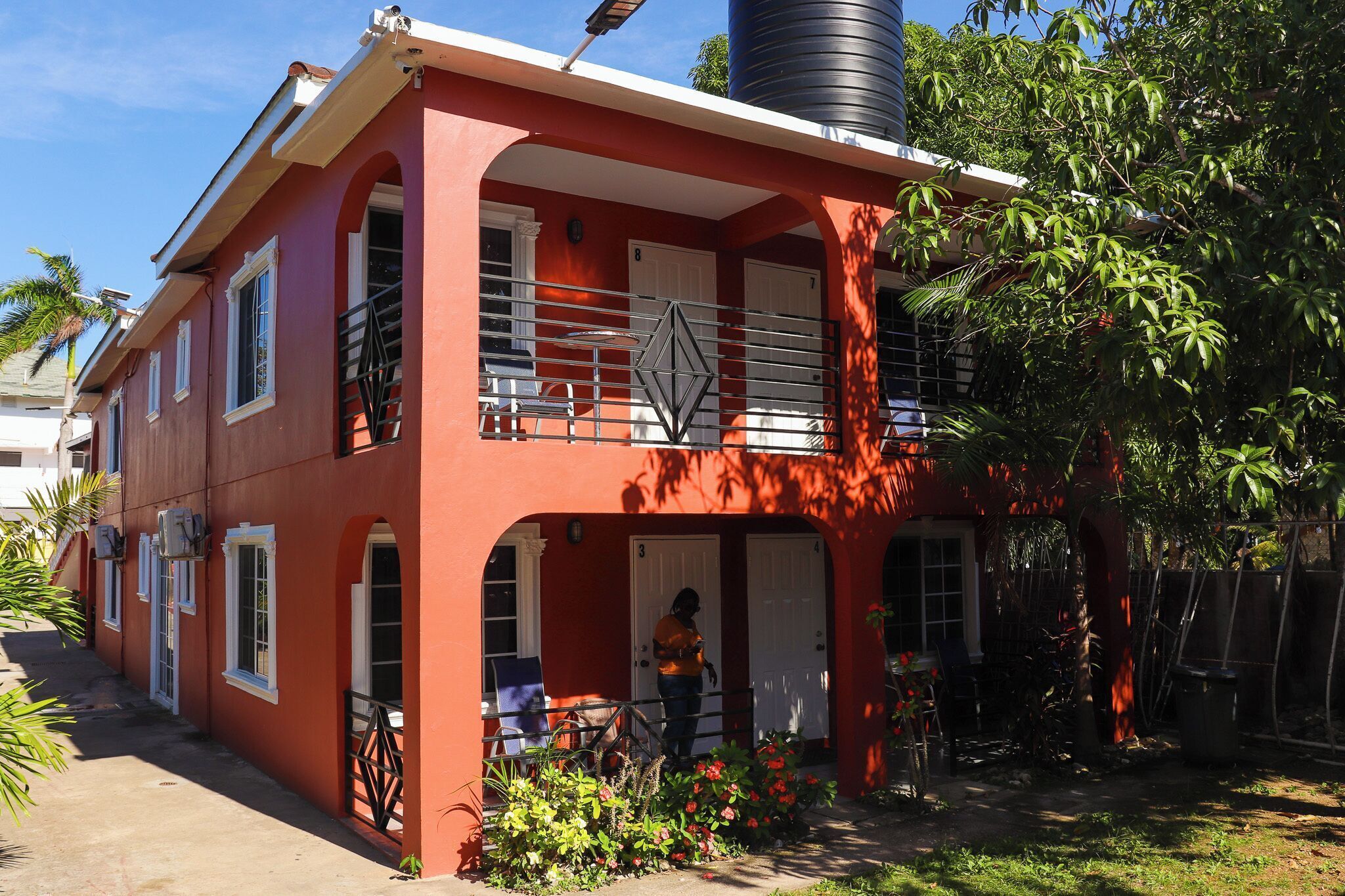 A red two-story house with a balcony, a water tank on the roof, and a garden with plants and flowers.