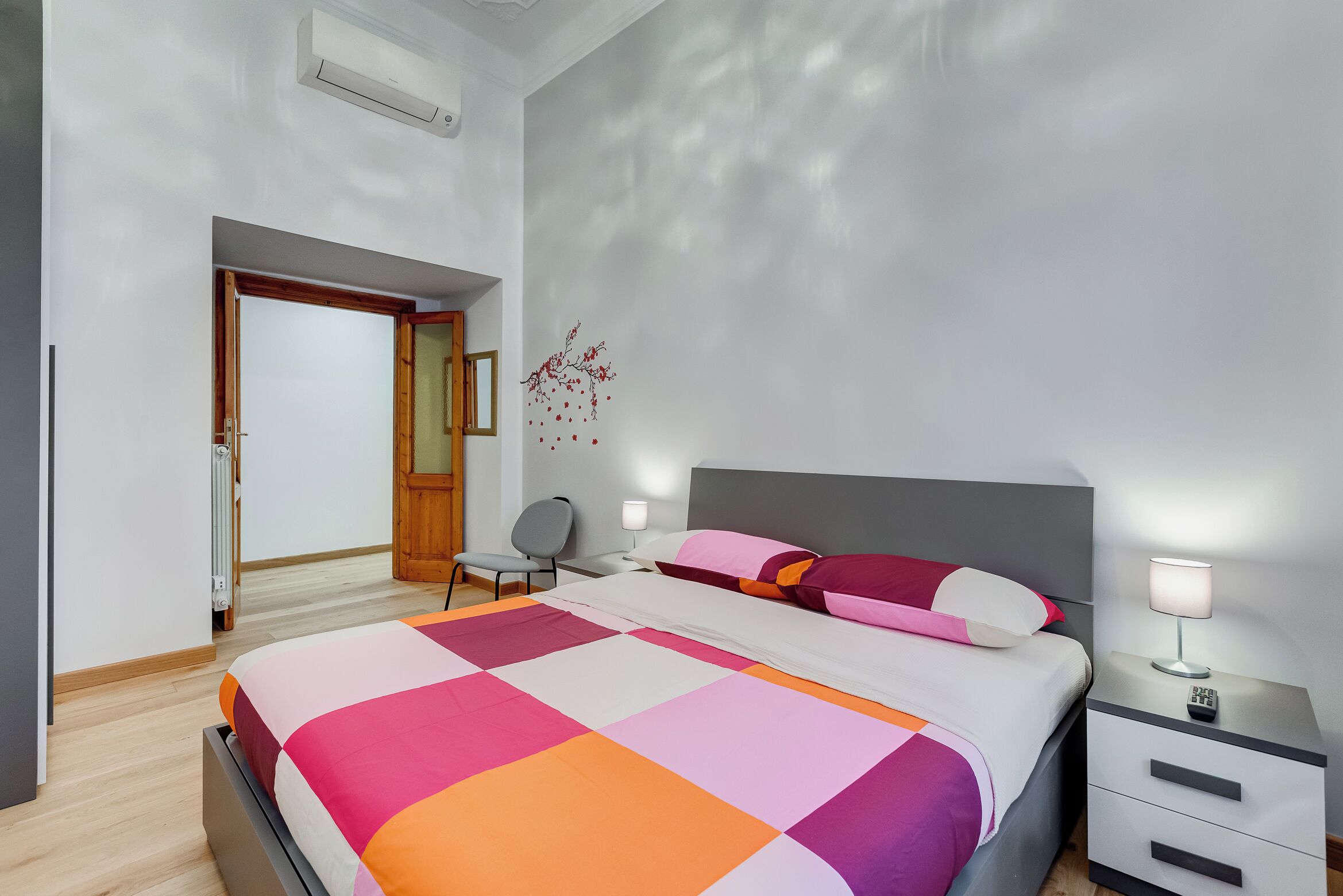 A modern bedroom with a large bed, bedside tables, a chair, and a door leading to another room.