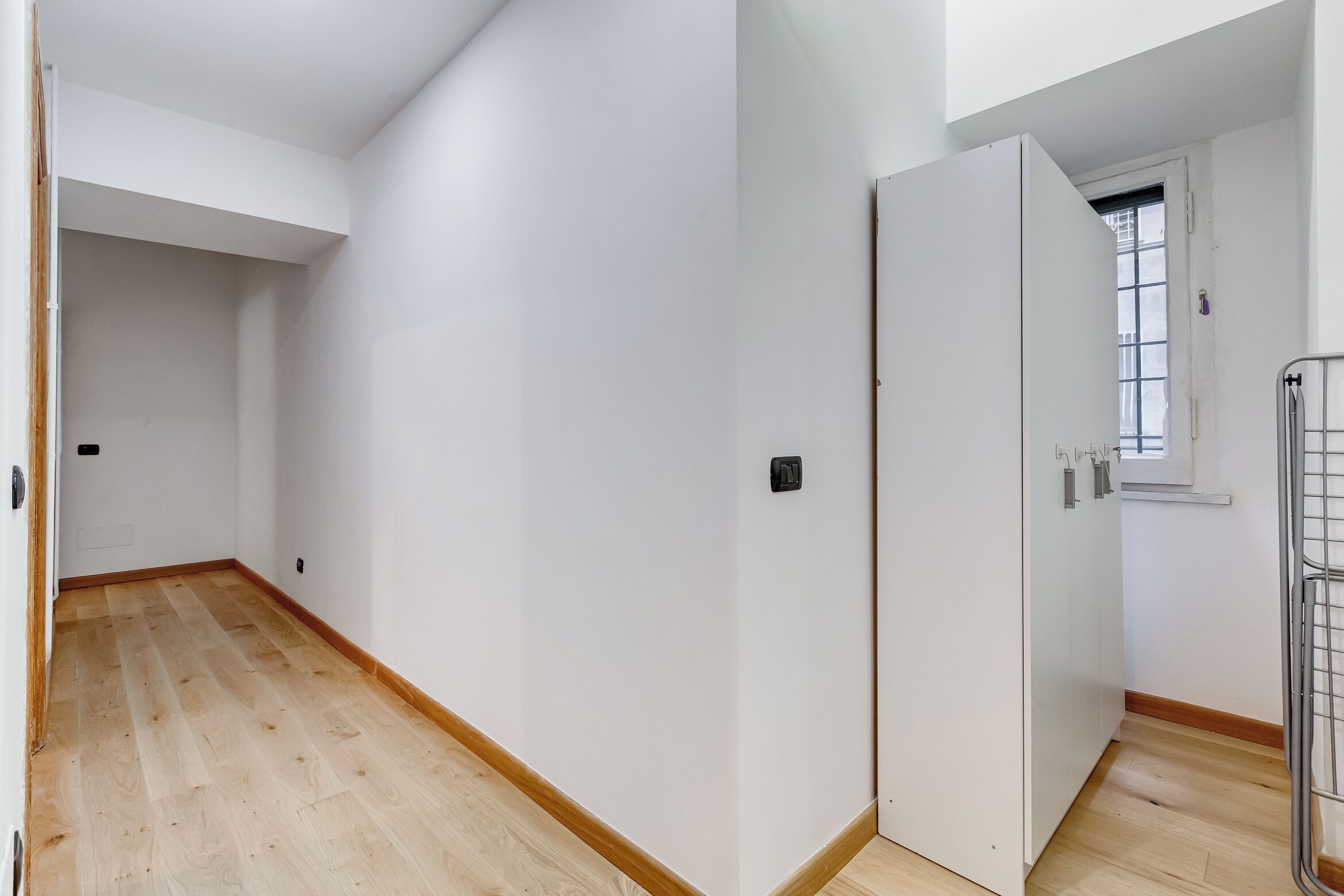 Un couloir étroit doté d’un plancher en bois et de murs blancs, avec une porte munie d’une fenêtre et une rambarde en métal sur le côté droit.