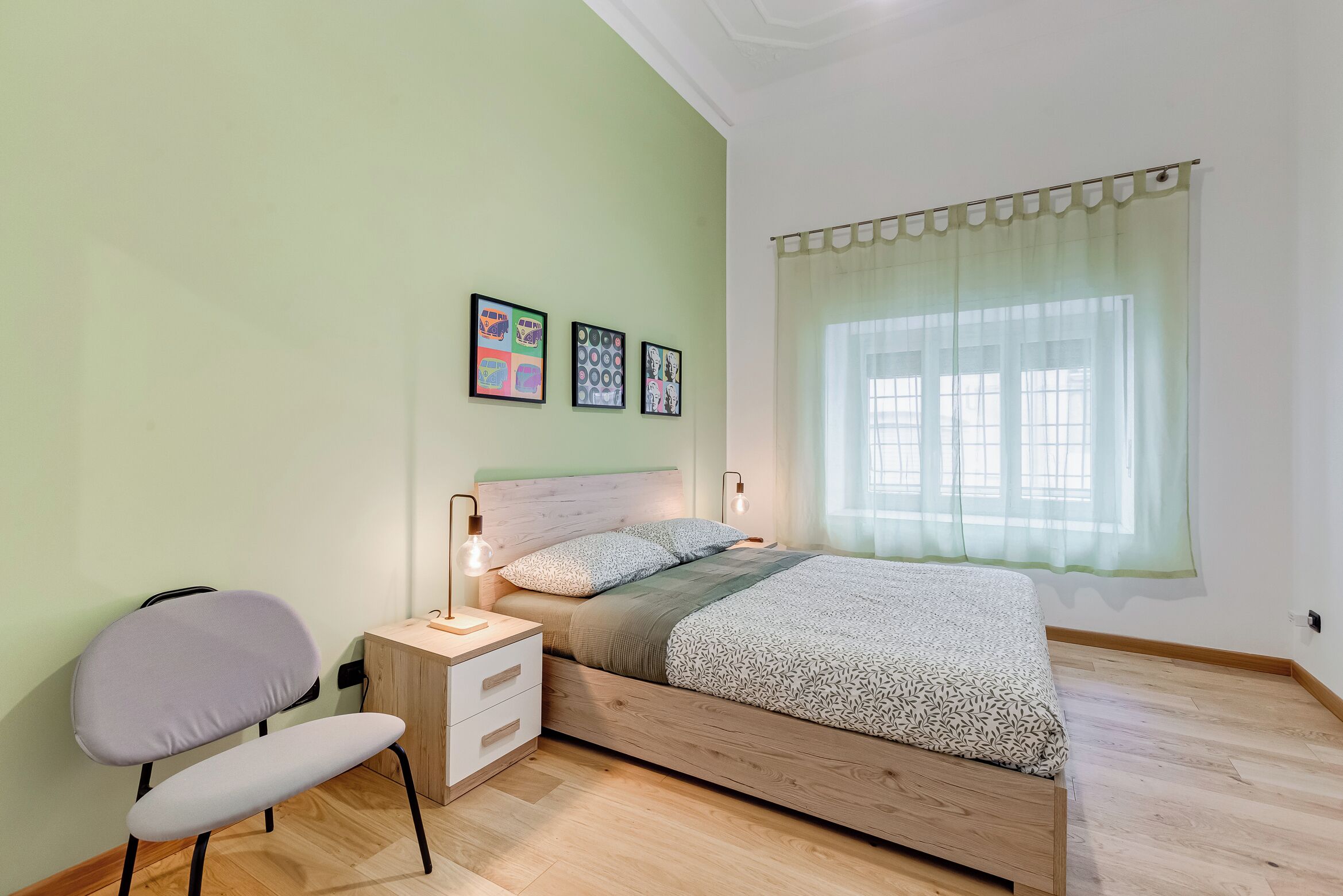 Ein Schlafzimmer mit einem hölzernen Bett, einem Nachttisch, einem Sessel und einem Fenster mit durchsichtigen Vorhängen.