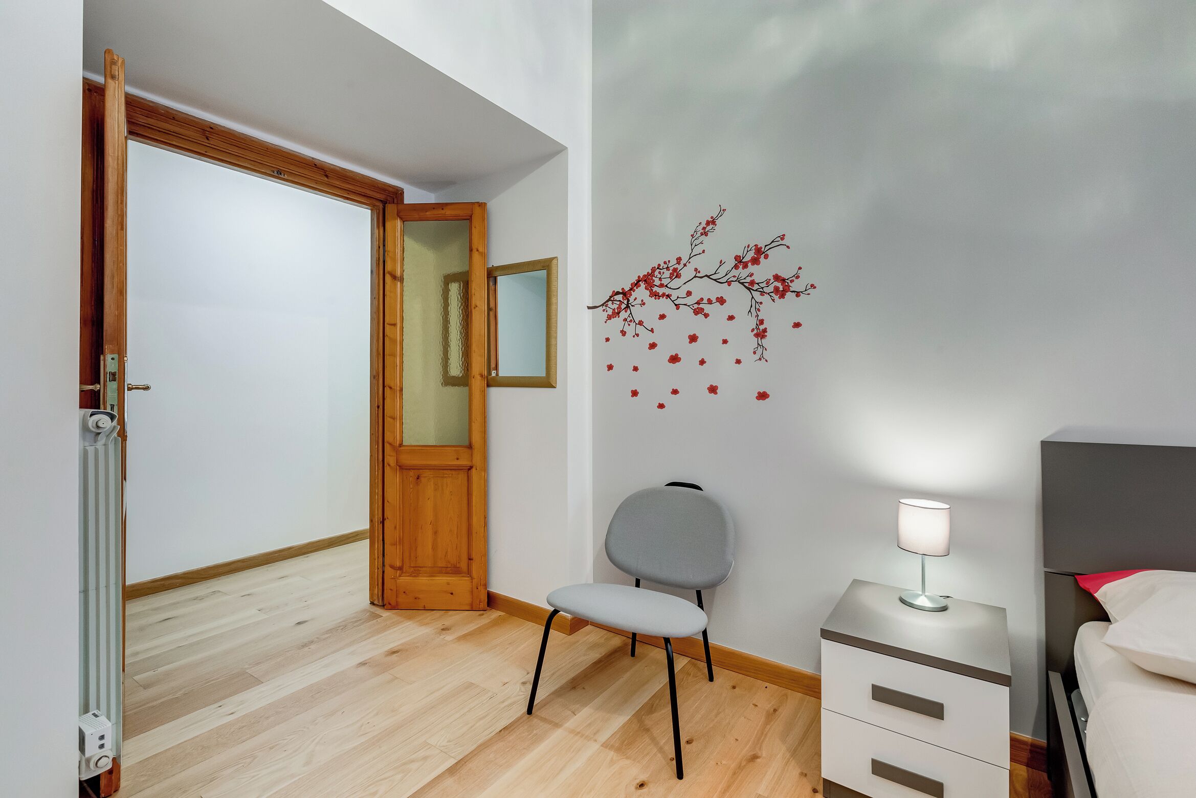 Un dormitorio con puerta de madera, una silla, una mesita de noche con lámpara y una cama.