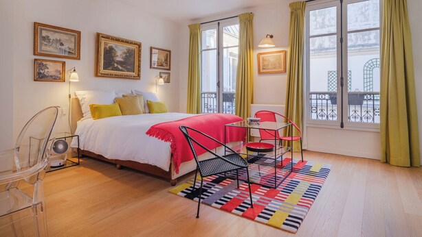 Un dormitorio con cama, una silla, una mesa y un ventanal con cortinas.