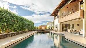 A swimming pool with a stone-lined edge, surrounded by lush greenery and a building with outdoor seating and a balcony.