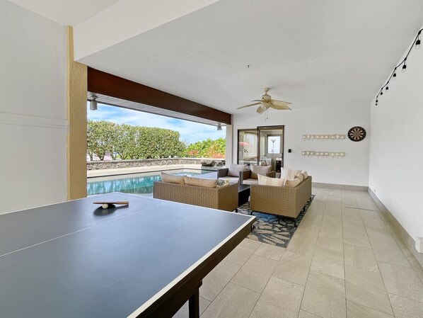 Una habitación con mesa de ping pong, muebles de mimbre, ventilador de techo y vista a una piscina.