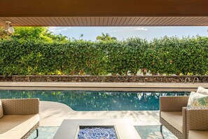 A poolside area with a stone wall, a clear pool, and wicker furniture. - Luxury Home w/Pool, King Suites & Sunset Views (Kailua-Kona)