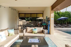 A poolside area with a sofa, a table, and a fire pit. There are umbrellas and lounge chairs in the background. - Luxury Home w/Pool, King Suites & Sunset Views (Kailua-Kona)