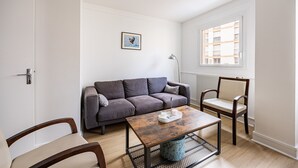 Ett modernt vardagsrum med en grå soffa, ett soffbord i trä, en blå fåtölj och ett fönster med utsikt över byggnader.