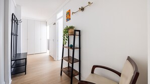 En hall med trägolv, en svart metallbänk, en trästol, en svart metallhylla med en växt och en väggmonterad lampa.