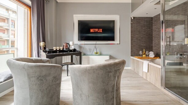 A modern living room with a flat-screen TV displaying the Netflix logo, a glass coffee table, two grey armchairs, a small side table, and a view of a balcony with a cityscape. - Four Seasons Suite in City Center, 350m from the Sea • 3 Suite - View & Balcony (Pescara)