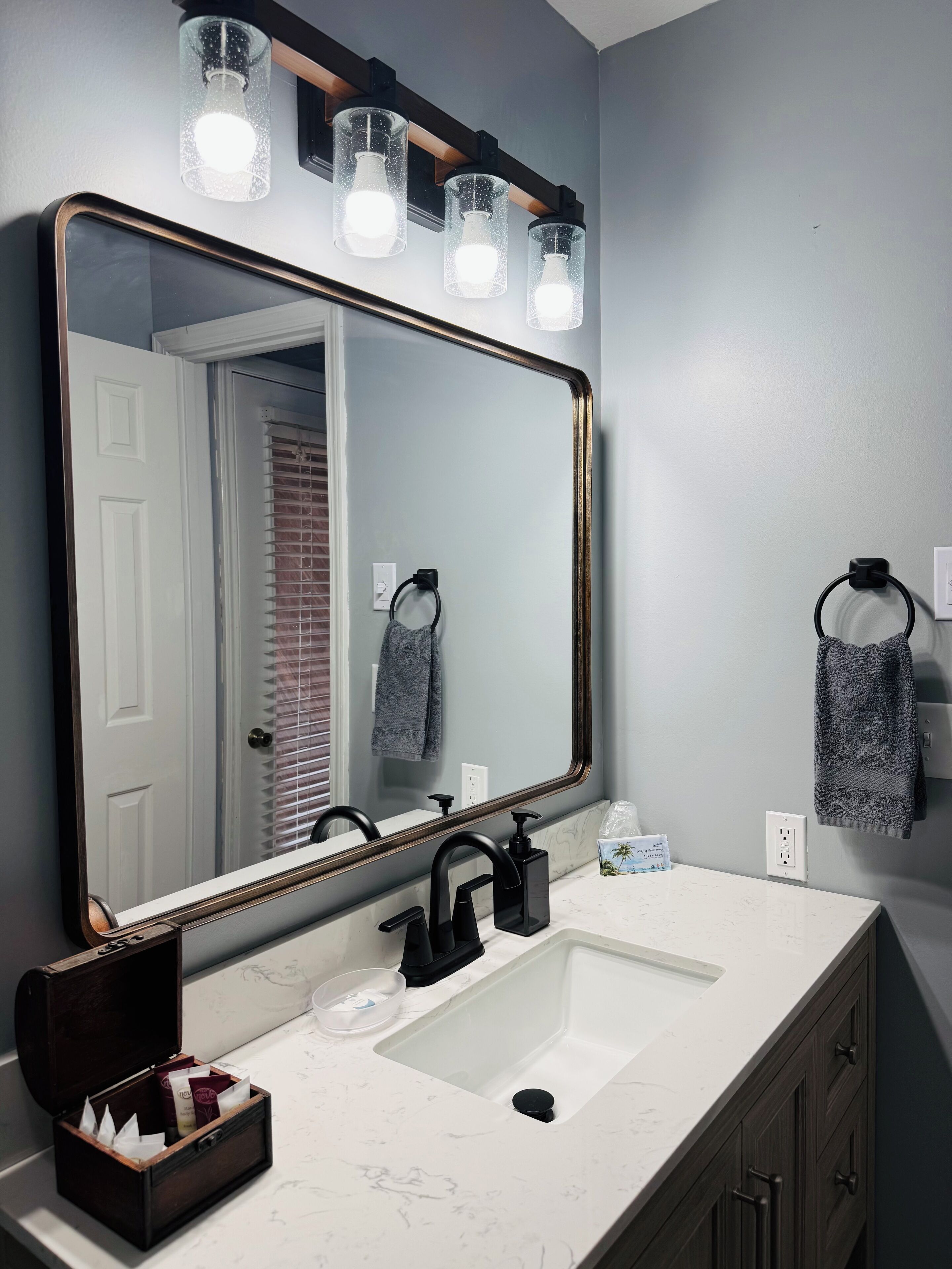 Un baño con un espejo grande, un lavabo blanco y un tocador con un armario de madera oscura. Hay tres lámparas sobre el espejo y un perchero en la pared.