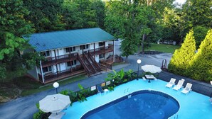 A swimming pool with lounge chairs, a covered seating area, and a building with a balcony in a wooded area. - Timber Pointe Resort (Jamestown)