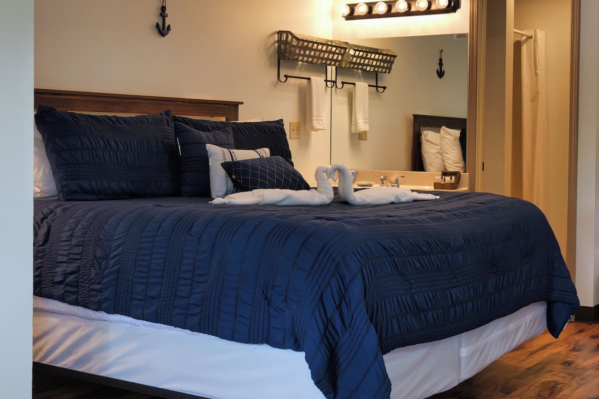 Un lit bien fait, avec une couverture bleu foncé et du linge de lit blanc, une tête de lit en bois, un luminaire fixé au mur et un miroir agrémenté d’un porte-serviettes.