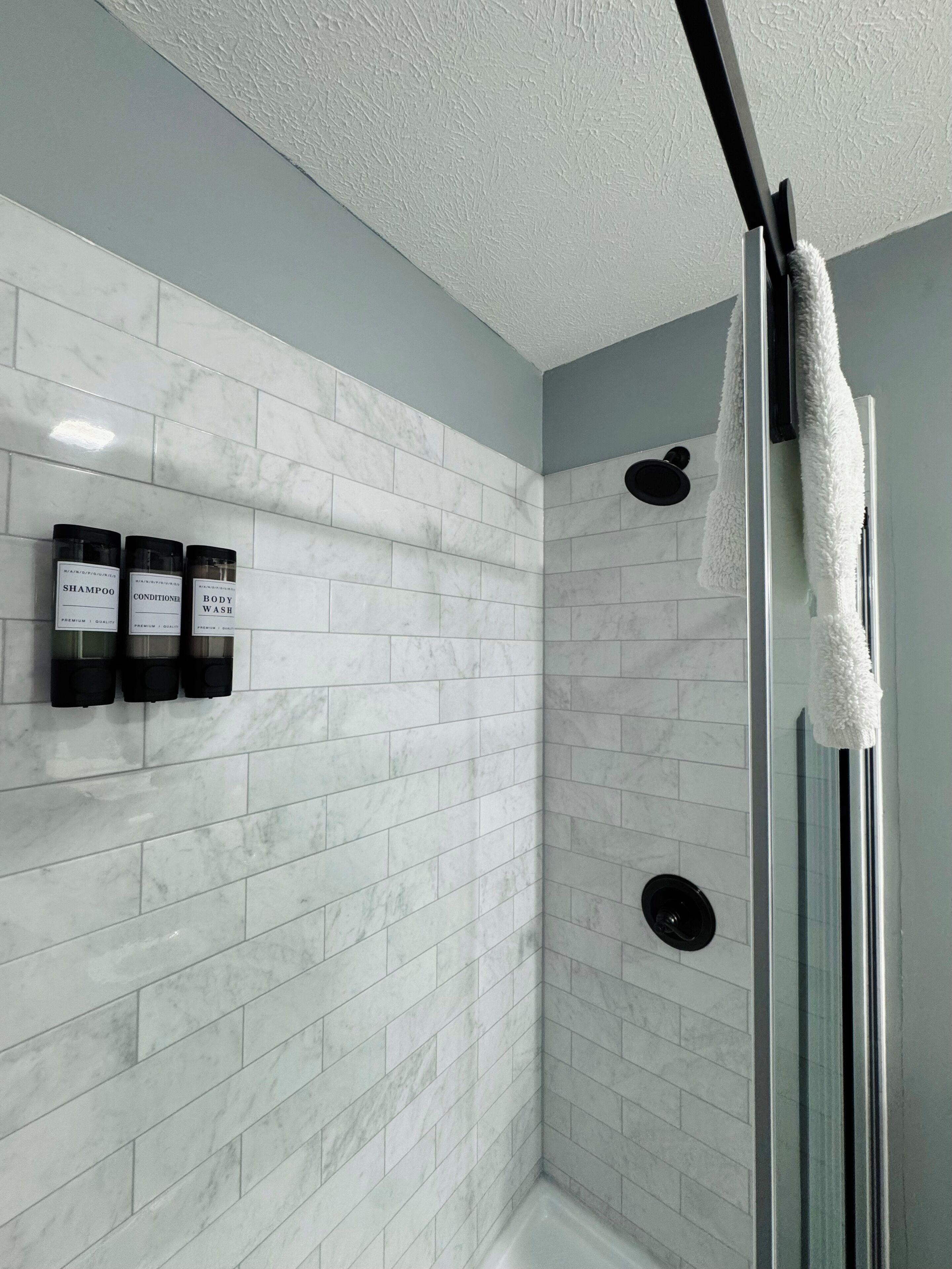 A modern bathroom with white marble tiles, a glass shower enclosure, and three bottles of shampoo, conditioner, and body wash on the wall.