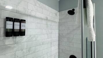 A modern bathroom with white marble tiles, a glass shower enclosure, and three bottles of shampoo, conditioner, and body wash on the wall.