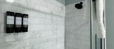 A modern bathroom with white marble tiles, a glass shower enclosure, and three bottles of shampoo, conditioner, and body wash on the wall.