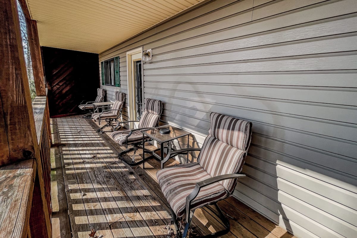 Une véranda couverte avec des chaises à rayures et une table, un plancher en bois et une vue sur une maison aux bardages blancs et aux volets verts.