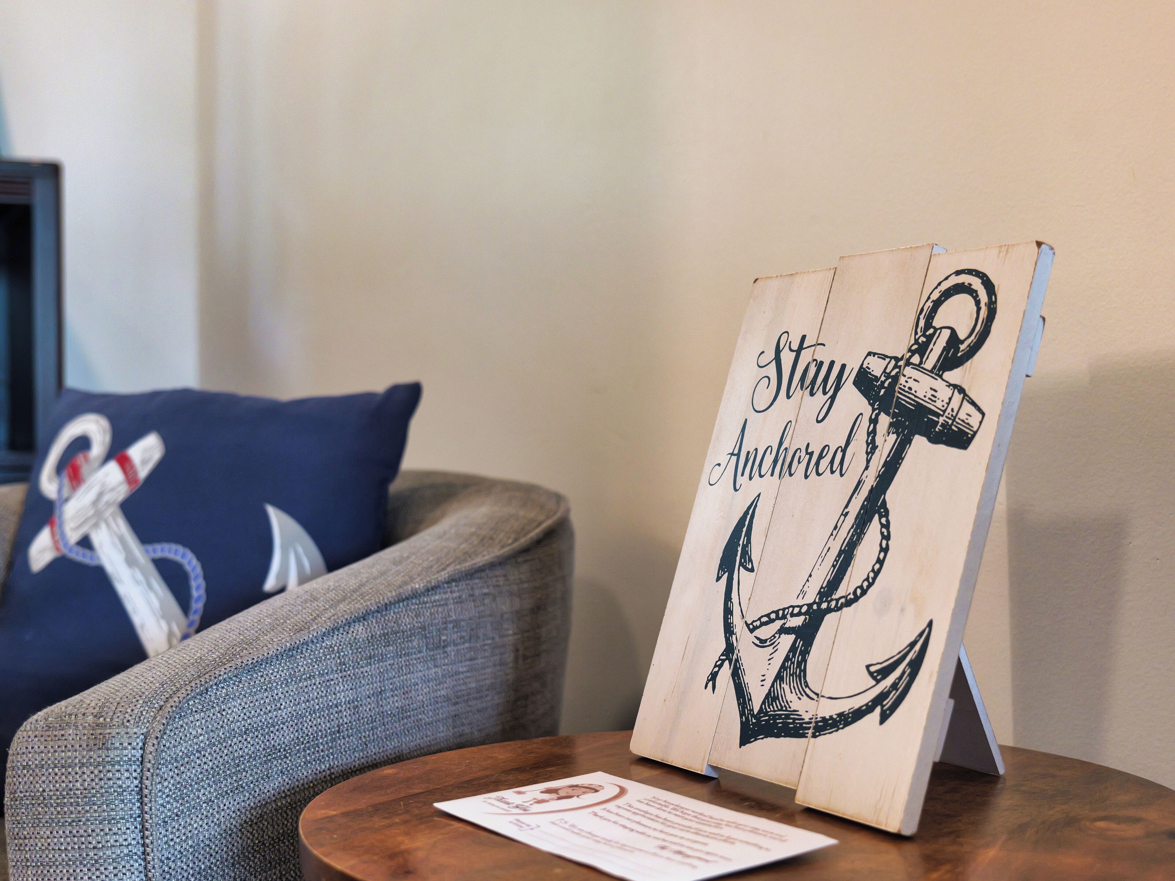 A wooden sign with an anchor and the text "Stay Anchored" on a table in a room with a grey sofa and a blue pillow with an anchor design.