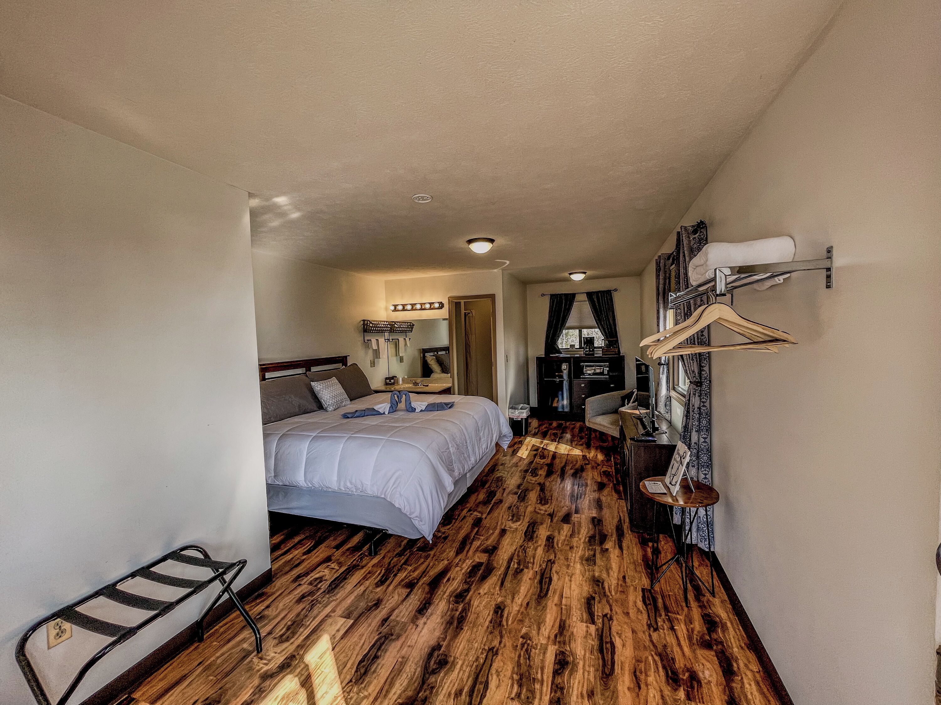 A compact hotel room with a bed, a desk, a chair, a mirror, and a coat rack.