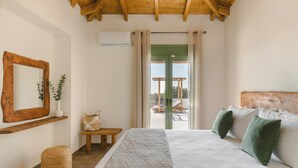 Un dormitorio con cama, cabecera de madera, espejo, aire acondicionado instalado en la pared, ventana con cortinas y vistas a una terraza exterior.