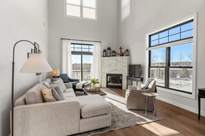 A spacious living room with a fireplace, large windows, and a high ceiling.