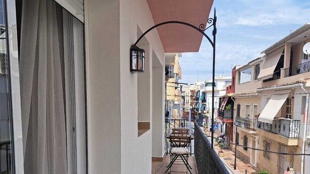 Un balcón con piso de madera, una mesa con sillas, una lámpara colgante y vistas a edificios vecinos.