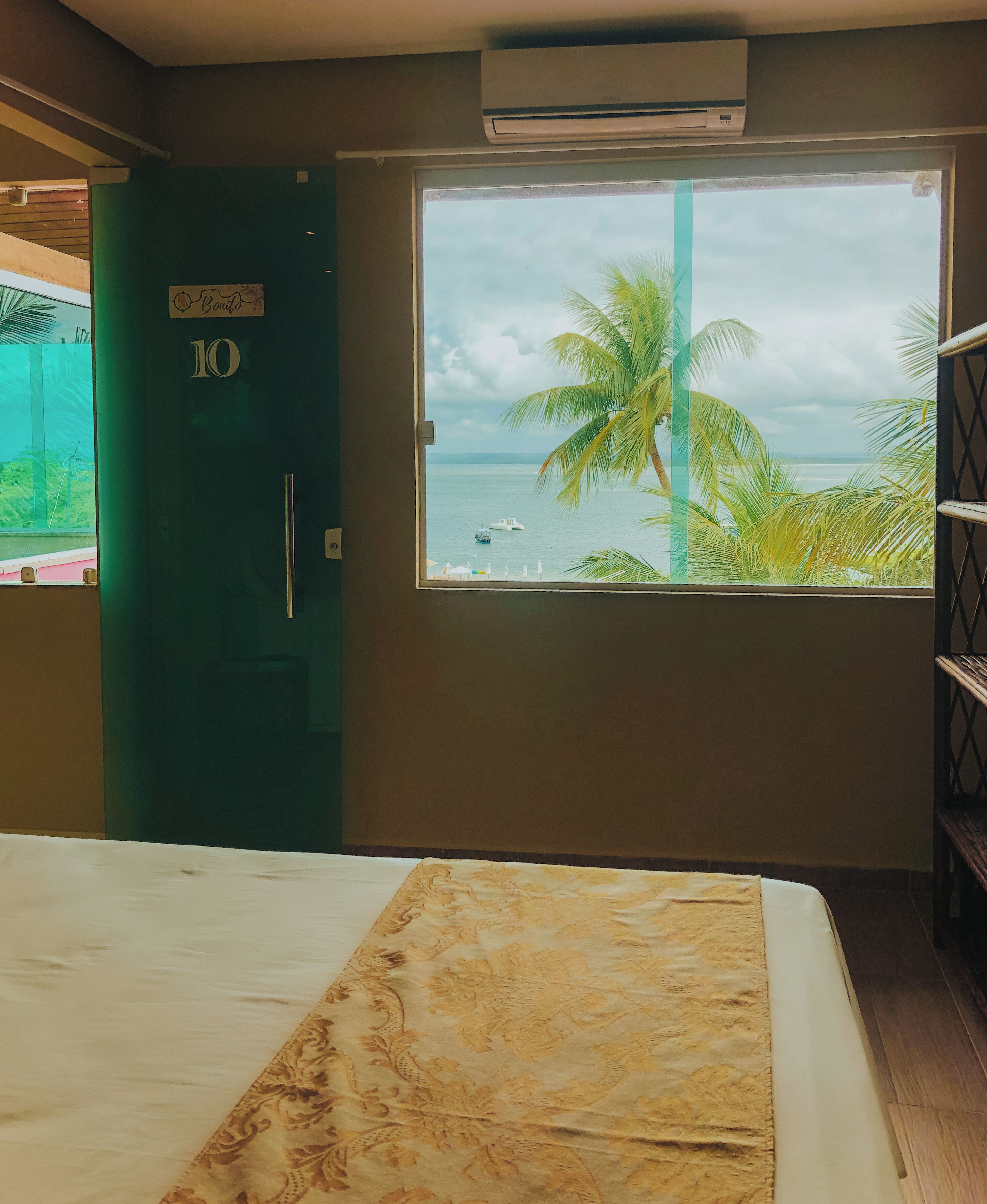 Une chambre avec un lit, une fenêtre donnant sur la mer et les palmiers, une porte portant l’inscription « 10 », et un appareil de climatisation.