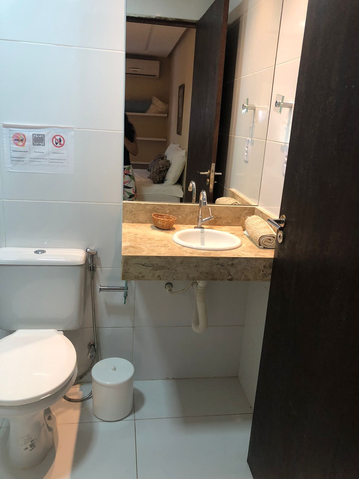 Un baño pequeño con inodoro, lavamanos y espejo. Hay un cartel en la pared con símbolos y texto.