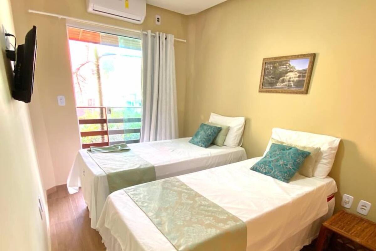 Une chambre d’hôtel avec deux lits simples, un plancher en bois, une fenêtre avec des rideaux, un appareil de climatisation et une petite table de chevet en bois.