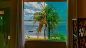 Une chambre dotée d’une grande fenêtre donnant sur une plage, un palmier et un bateau sur l’eau.
