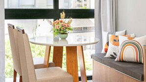A modern living room with a round glass table, beige chairs, a sofa with colorful cushions, a hanging plant, and a view of tall buildings through large windows. - Gastronomy | Pool | 500m from Beiramar #CA76 (Florianópolis)