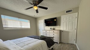 Een slaapkamer met een bed, een plafondventilator, een televisie, een dressoir en een raam met jaloezieën.