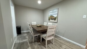 Een eethoek met een houten tafel en stoelen, een prullenbak en een schilderij aan de muur.