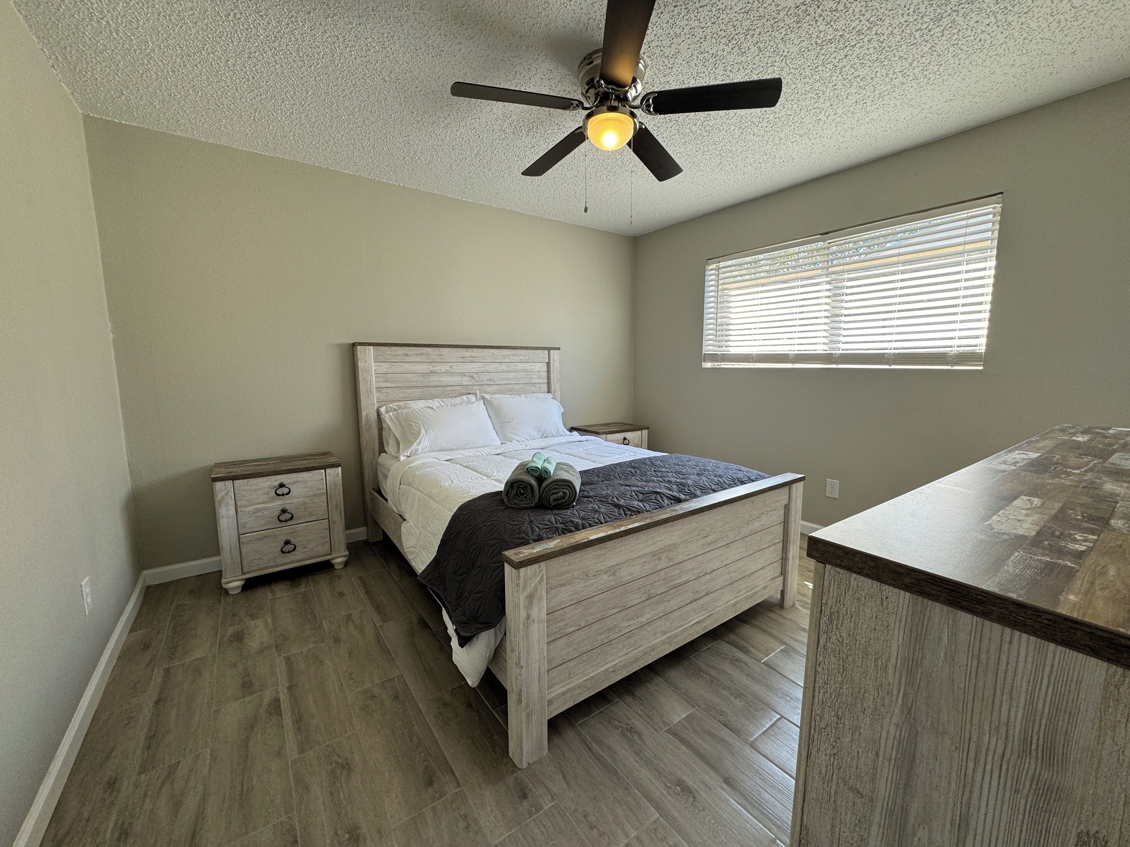 A bedroom with a bed, a nightstand, a ceiling fan, a window with blinds, and a wooden floor.