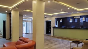 Un vestíbulo de hotel con un mostrador de recepción, una zona de estar con sillones naranjas y una escalera que sube a otro piso.