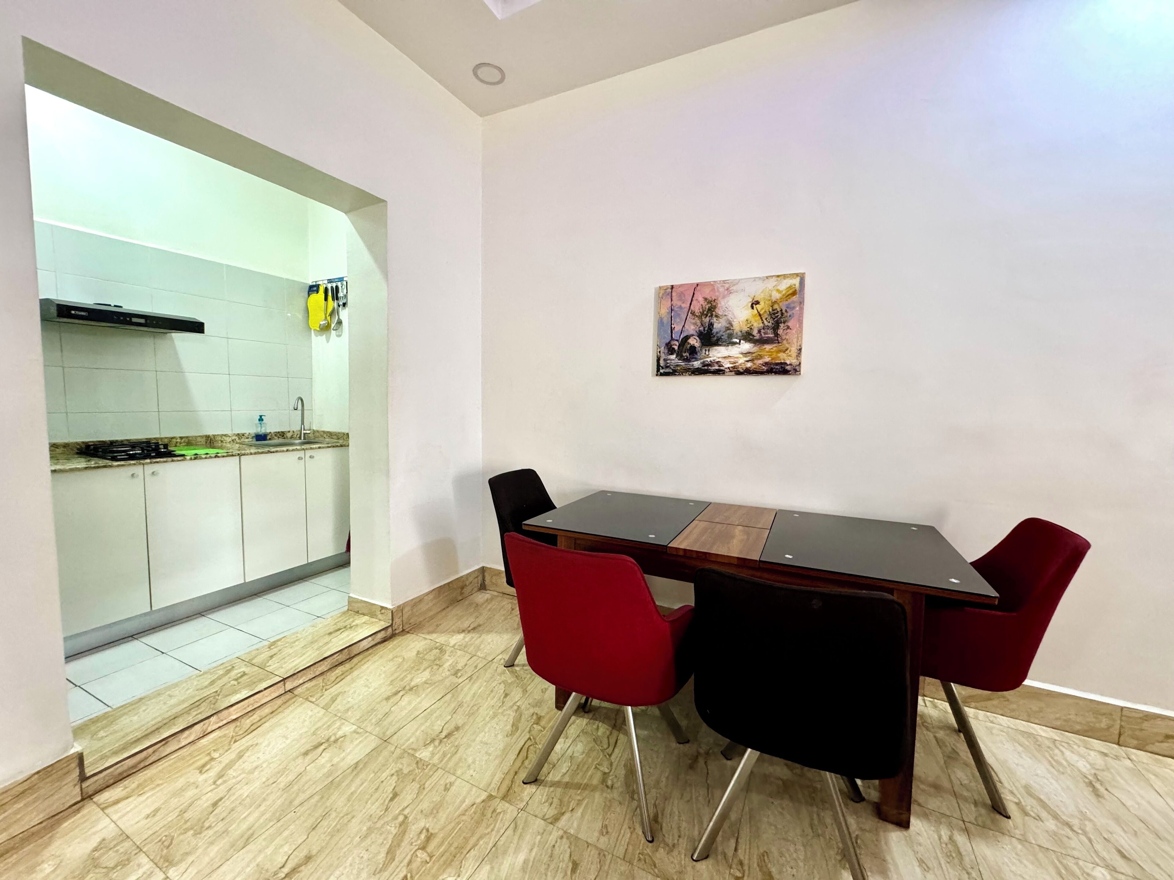 A dining area with a wooden table and red chairs, a kitchen with white cabinets, and a painting on the wall.