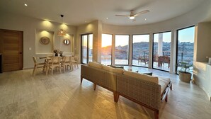 A spacious living area with a large sofa, dining table, and chairs, a ceiling fan, and large windows offering a view of the outdoors. - Desert Retreat: Heated Salt Water Plunge Pool* Grill, Views, Cinema,nearBalandra (La Paz)