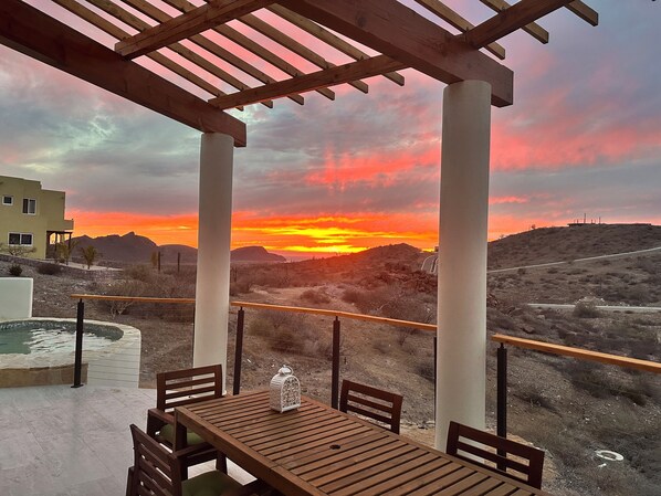 Una pérgola de madera con mesa y sillas, con vistas a un paisaje desértico al atardecer.